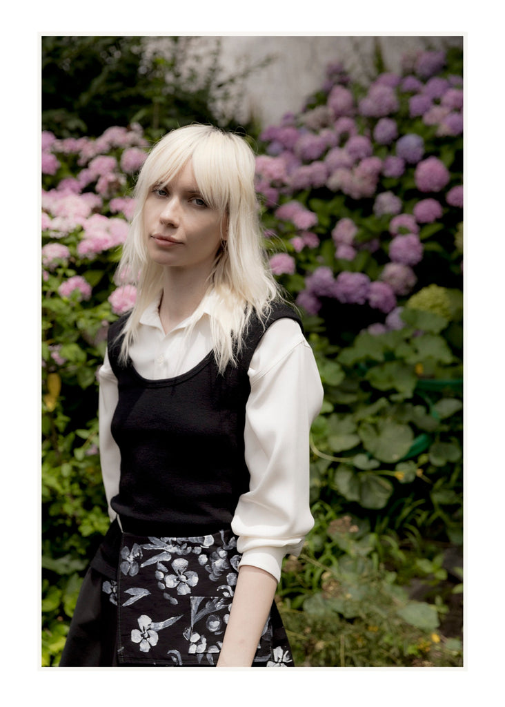 Woman with blonde hair wearing a black top and white shirt in front of purple flowers