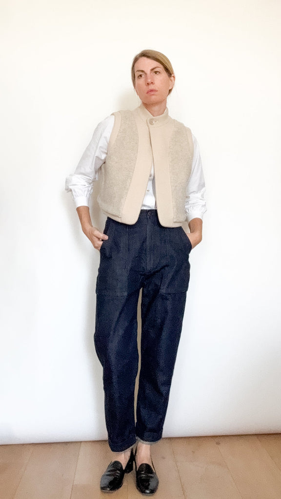 Person wearing a beige vest over a white shirt with dark pants against a white wall.