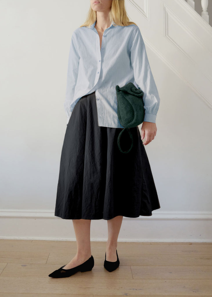 Person wearing a light blue shirt and black skirt holding a green bag indoors.