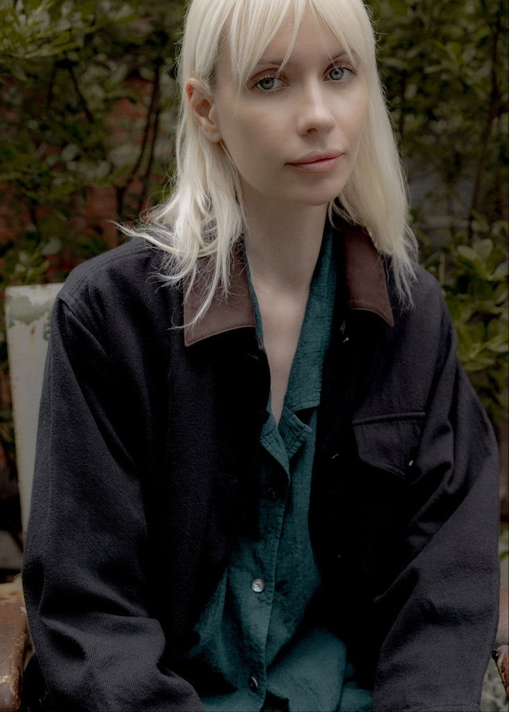 Person with blonde hair wearing a dark jacket over a green shirt, sitting outdoors.