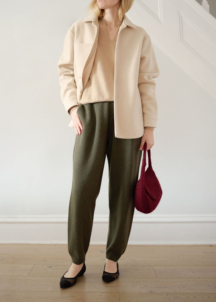 Woman wearing a beige jacket, beige shirt, green pants, and black shoes, holding a red bag indoors.