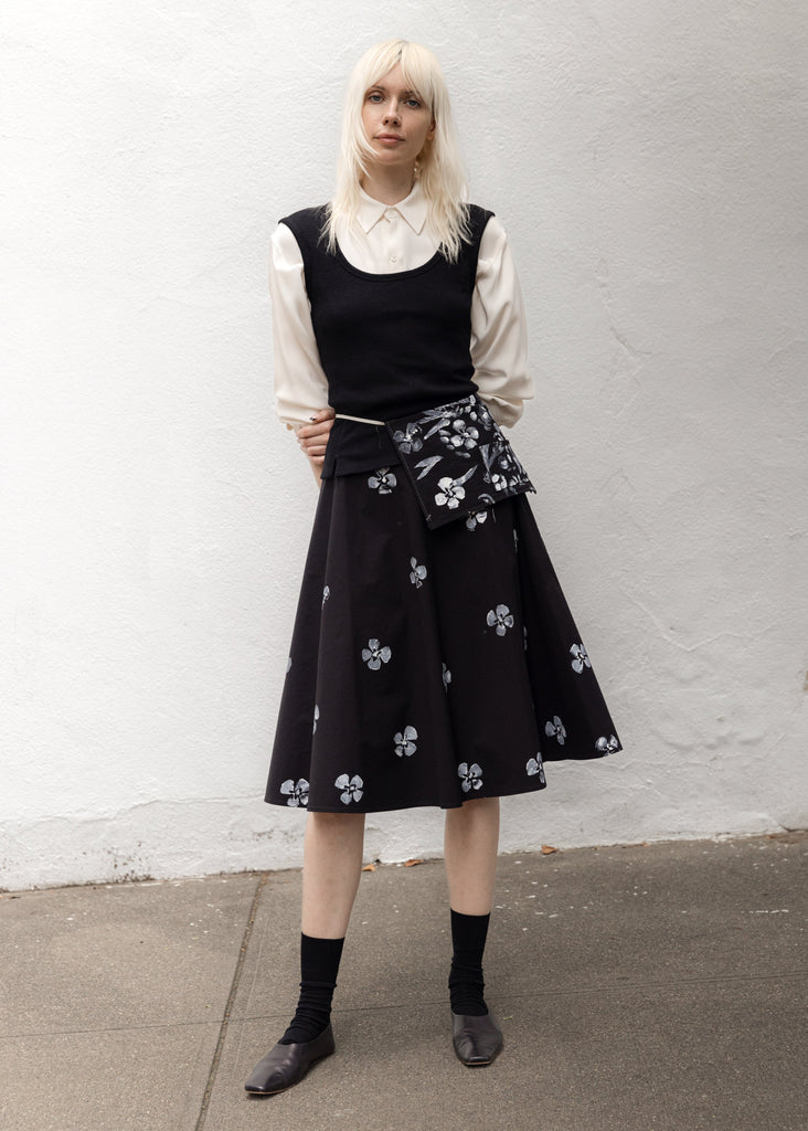 Person wearing a black sleeveless top and floral skirt against a white wall.