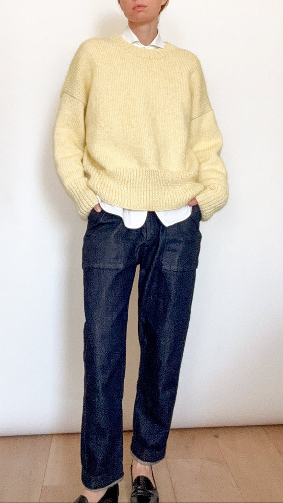Person wearing a light yellow sweater and dark blue jeans against a white wall.