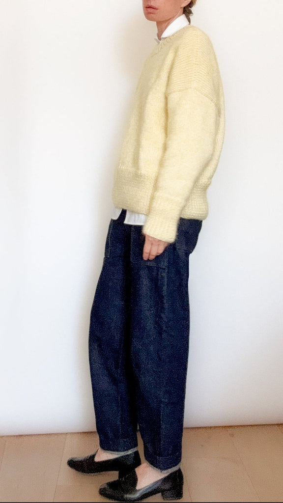 Person wearing a light yellow sweater and dark blue pants against a white wall.